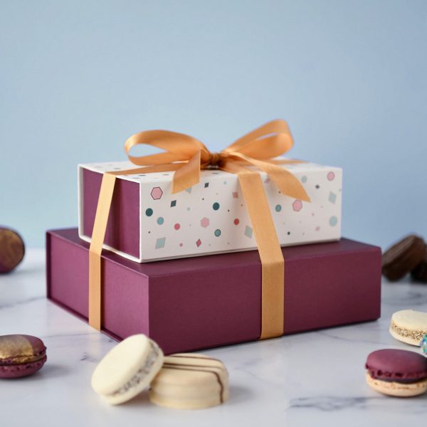 An Argentinian alfajores box of 12 on top of a box of 18 French macarons with a golden ribbon holding them together, and some assorted French macarons and alfajores lying around.