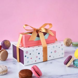 A macaron box of 3 on top of a box of 9 with a golden ribbon holding them together, and some assorted French macarons lying around.