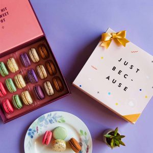 A purple background features a Woops! box of 18 French macarons with assorted flavors and a light pink sleeve with black letters at its center plus a golden ribbon. At the bottom is a white plate with French macarons and a little plant.