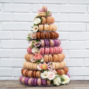 A large French macaron pyramid with assorted flavors is decorated with different types of flowers.