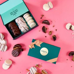 A pink background features an open green Woops! French macarons box full of assorted alfajores. Another green Woops! French macarons box with a golden ribbon and several alfajores lying around.