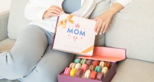 A woman is holding a Woops! French macaron box with a Mother’s Day sleeve in her hands. To her right is a box full of assorted macarons. 