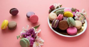 A plate full of assorted French macarons is surrounded by assorted macarons and flowers. 