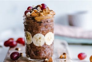 Attribute: A jar full of chocolate overnight oats, bananas, peanut butter, berries, and nuts is on top of a wooden board. Surrounding it are crushed nuts and berries