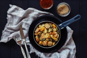 A pan is full of roasted vegetables and chicken. Surrounding it is a fork and knife plus some spices. 