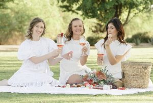 Three women sharing a picnic blanket are eating French macarons and holding champagne flutes in their hands.