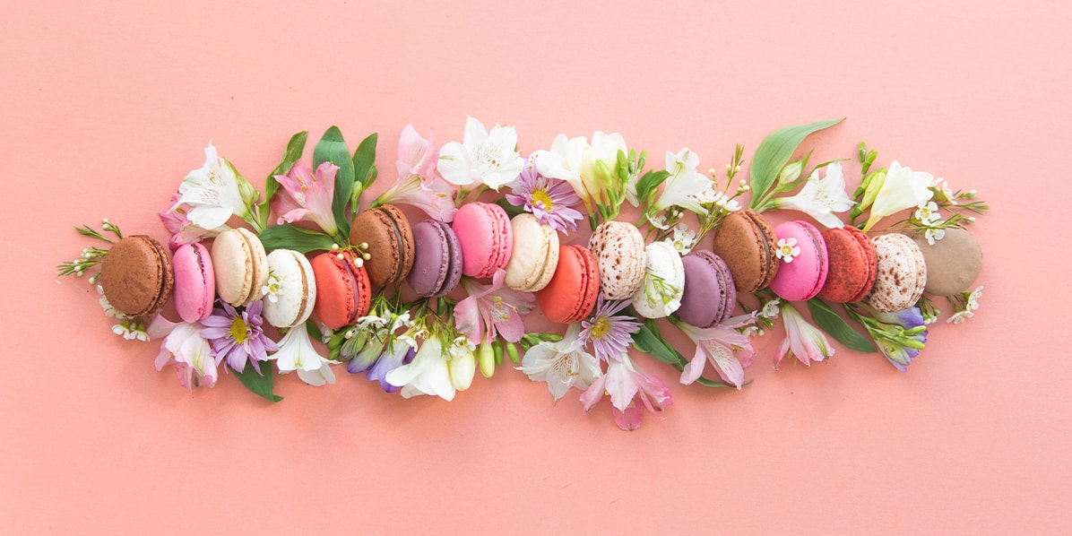 Numerous assorted French macarons are in a horizontal line. Behind them are numerous flowers.