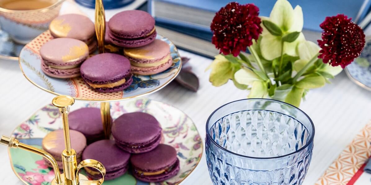A tier tray full of Honey Lavender French macarons is surrounded by blue cutlery, books, and flower vases.