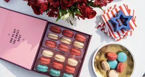 A tray full of red, white, and blue French macarons has some golden stars on top. To its side is a box of French macarons with a light blue sleeve and to the right several macaron favor boxes of two macarons