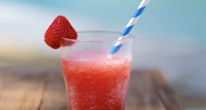  A glass full of a pink daiquiri cocktail has a strawberry on one of its borders and a blue and white straw