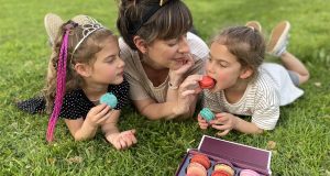A mom and her two girls are lying on the grass eating French macarons. In front of them is a Woops! Macaron box of 9