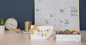 A box full of French macarons has a Woops! Macaron box with a Thank You sleeve to the left. Behind them are several office implements.