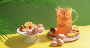 Numerous Strawberry Lemonade French macarons are surrounded by lemons and strawberries. A big jar of strawberry lemonade stands to the right