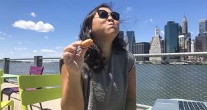 A woman is holding a Woops! French macaron. Behind her is the New York skyline. 