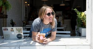 A smiling woman is holding a Woops! Macarons & Gifts cup full of coffee