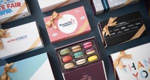 A box full of nine assorted French macarons is surrounded by macaron boxes with different sleeves. 