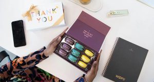Two female hands are holding a box full of nine French macarons. To the left is a Woops! Box of macarons with a Thank You sleeve and lying around are several office implements. 