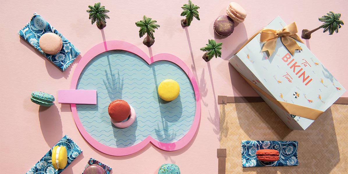 Several macarons are in a fake pool and tanning. A French macaron box with a Summer sleeve stands to the right.