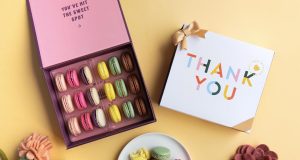 A box full of assorted French macarons has a macarons box with a Thank You sleeve to the right. Below is a plate full of macarons and some flowers.
