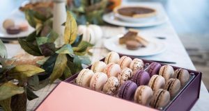 A box full of assorted French macarons is on top of a table with plant-decorated candles and other plates with macarons and assorted sweets