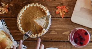 Two female hands are cutting a pumpkin pie. Lying around are some fall leaves, a wooden table, and a cup full of red liquid