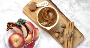 A plate full of apples has a wooden board with pumpkin butter, cinnamon sticks, hazelnuts, and a spoon with nutmeg to the right.
