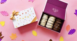 A box full of 12 assorted alfajores has a French macaron box with a Thanksgiving sleeve to the left. Lying around are numerous paper leave