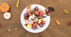 A plate full of assorted macarons is surrounded by small pumpkins and fall leaves. 