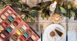 A box full of assorted macarons is surrounded by plates with assorted desserts and fall decorations. 