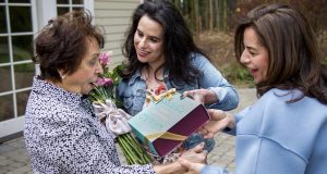 Three smiling women have a bouquet of flowers and a box of macarons between them