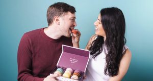 A smiling woman is giving a macaron to the man on the left. He’s holding a box of French macarons