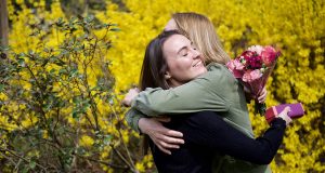 Two women are hugging. One of them is holding a bouquet of flowers and a box of macarons