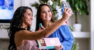 Two smiling women are taking a selfie while holding a box of macarons. Behind them is a Christmas tree.