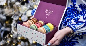 Two female hands are holding a box of French macarons. Behind her is a Christmas tree