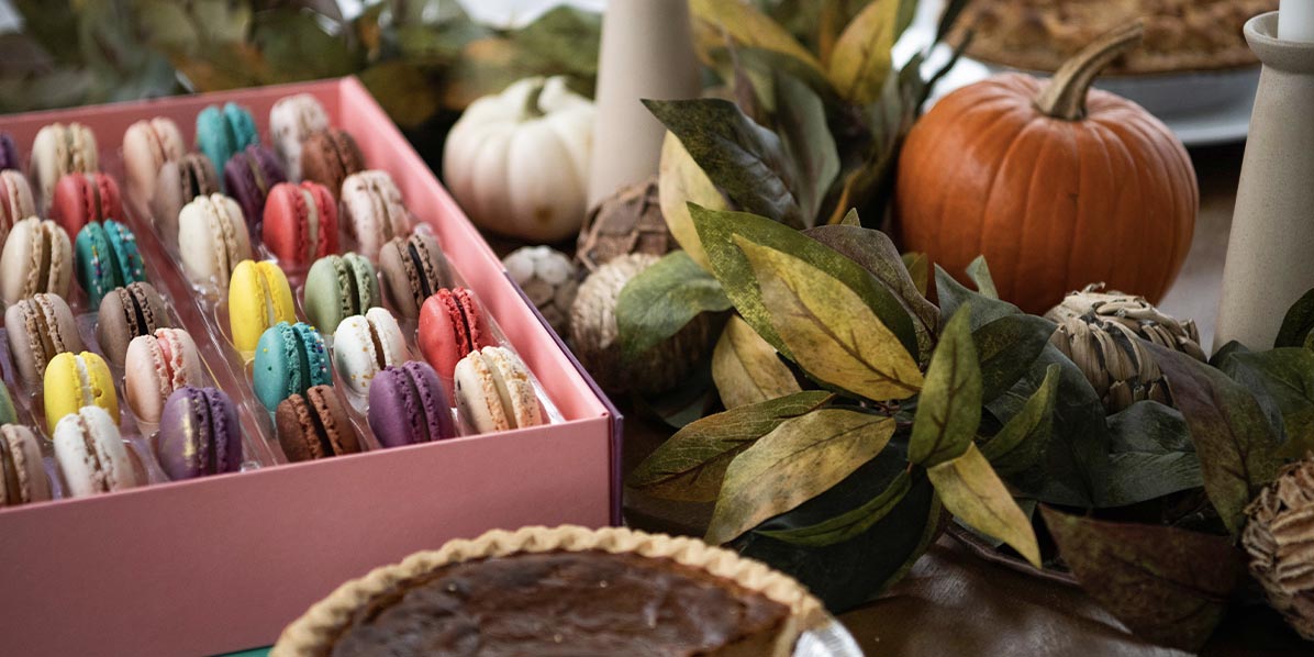 A box full of macarons and a plate with three French macarons are on top of a table decorated with fall ornaments and surrounded by other assorted desserts