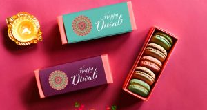 A box full of five assorted macarons is surrounded by French macaron favor boxes with Diwali sweets sleeves and Diwali candles. On the left is a candle