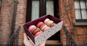 A male hand is holding a French macaron box in front of a doorstep.