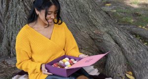A woman with a yellow sweater is sitting on a tree. On her lap is a box of macarons. 
