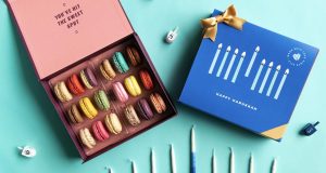 A box full of French macarons has a macaron box with a blue Hanukkah sleeve to the right. At the bottom are some candles