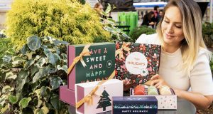 A smiling woman is sitting to the side of several French macaron boxes with Holiday sleeves. 