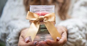 Two female hands are holding a French macaron favor box tied with a golden ribbon, inside are two macarons