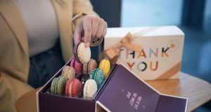 A woman is grabbing a macaron from a box full of macarons. To the right is a box of macarons with a Thank You sleeve. 