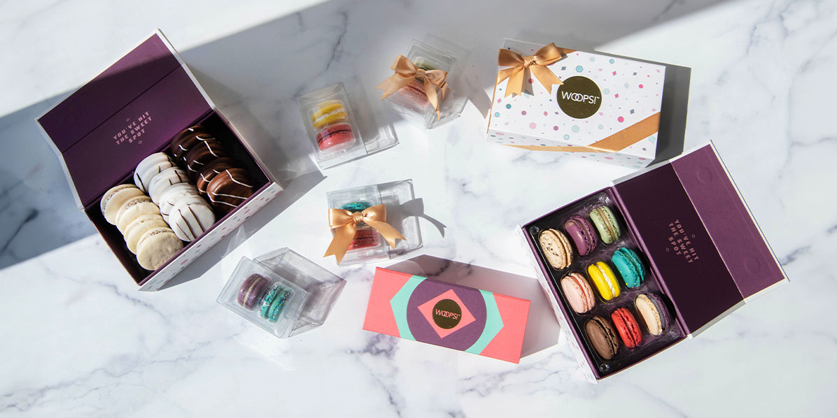 A box full of assorted macarons is surrounded by French macaron boxes and a box full of alfajores on top of a marble background.