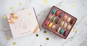 A box full of 18 assorted French macarons has a macaron box with a light pink New Year’s sleeve to the left. Surrounding them is golden confetti.
