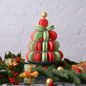 A French macaron pyramid with Red Velvet and Pistachio macarons is surrounded by holiday macaron boxes and Christmas ornaments