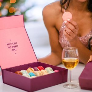 A woman holding a French macaron has two macaron boxes and a wine in front of her. Behind her are Christmas ornaments.