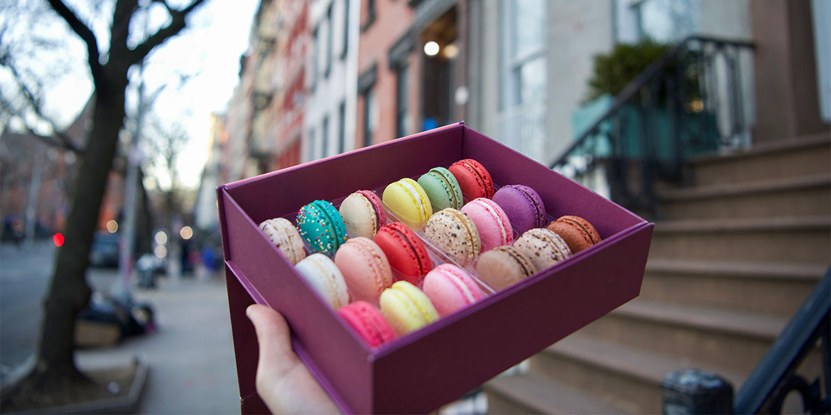 A hand is holding a box full of 18 assorted French macarons in front of a typical NYC doorstep.