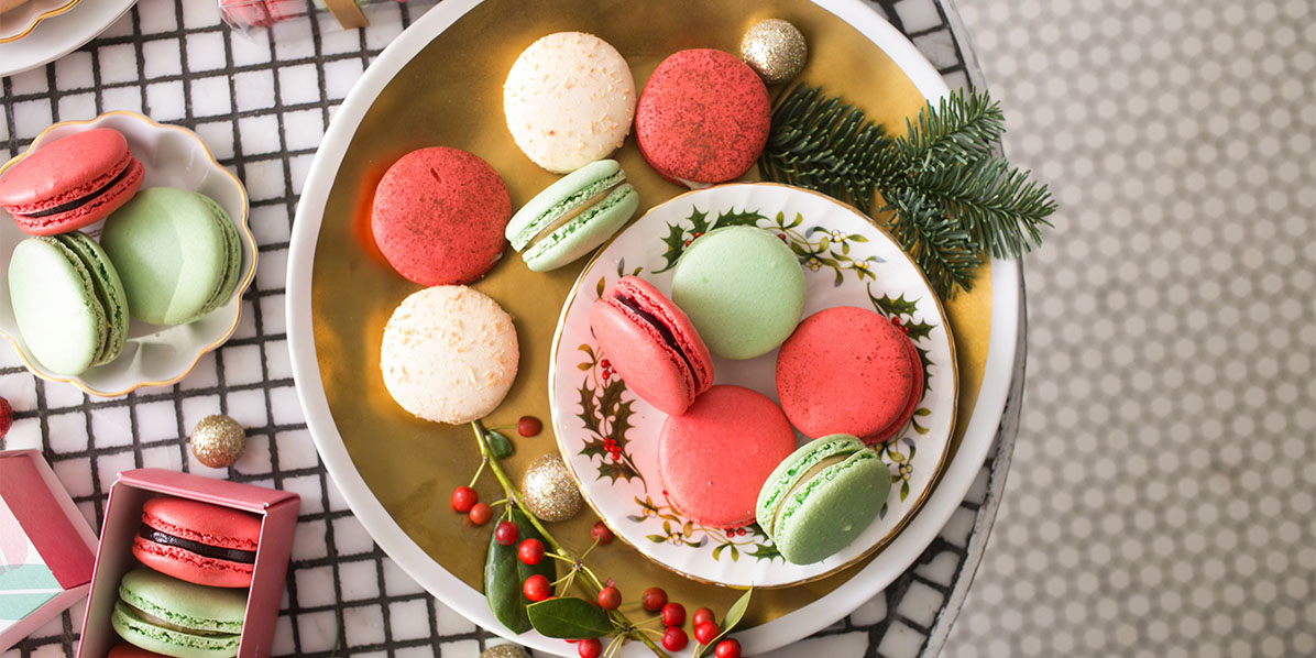 A plate full of Red Velvet and Pistachio Christmas macarons and Christmas ornaments is surrounded by macaron boxes and assorted macarons.
