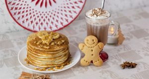 A plate full of pancakes has a gingerbread cookie and milkshake to the right. Behind is a red and while plate