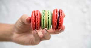 A hand is holding two Red Velvet macarons and a Pistachio one.